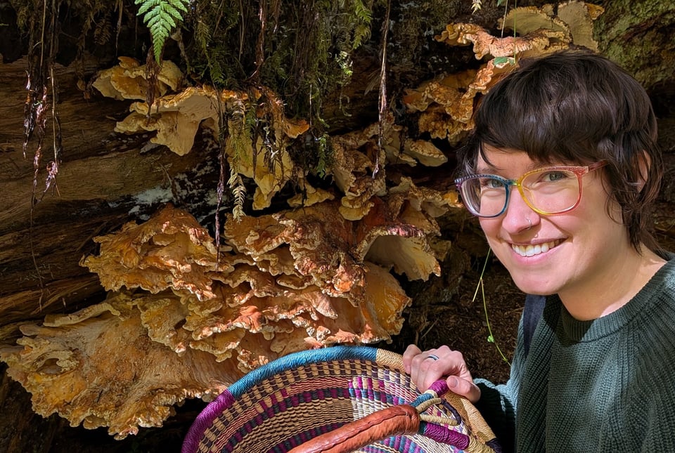Maia holds a basket and grins next to a huge flush of chicken of the woods growing out of a dead tree