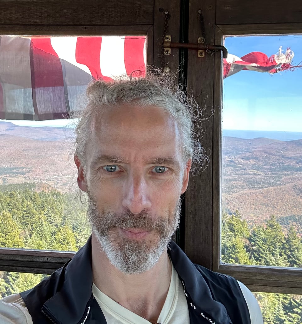 Selfie of white man in fire tower overlooking Catskills mountains; a slightly tattered American flag is fluttering behind him