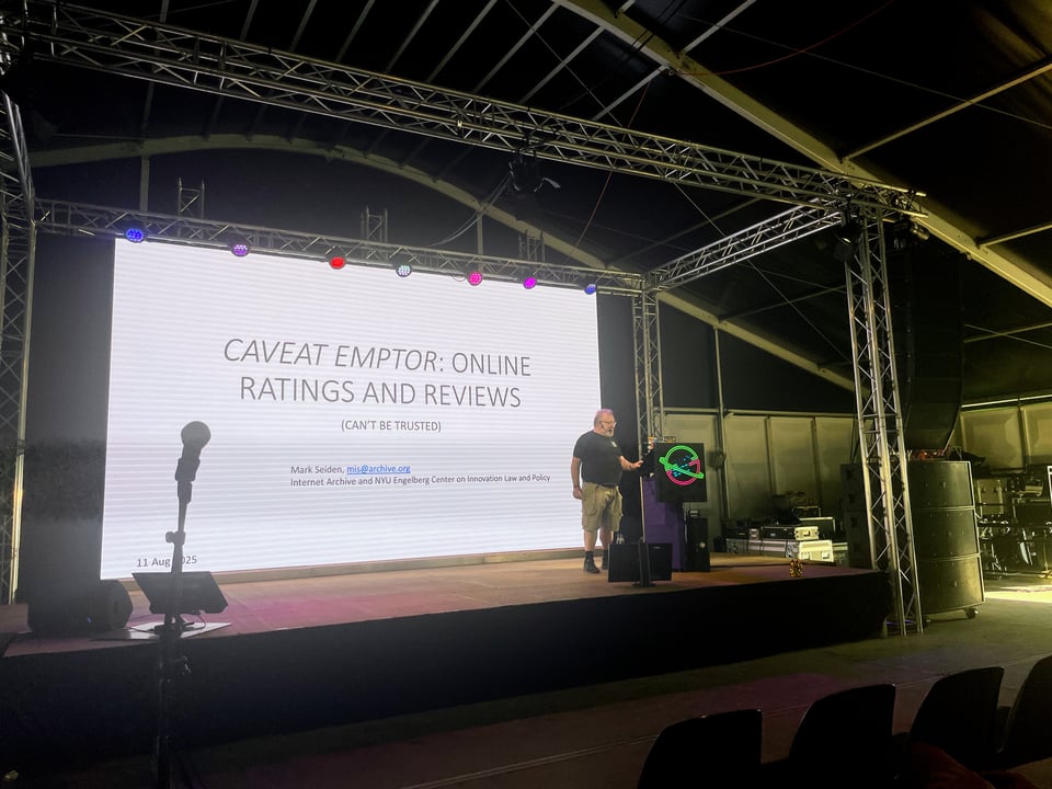 A man with an Internet Archive t-shirt speaks on stage with a WHY2025 logo on the podium.  The screen behind him reads "Caveat Emptor: Online Ratings and Reviews (Can't be trusted)"