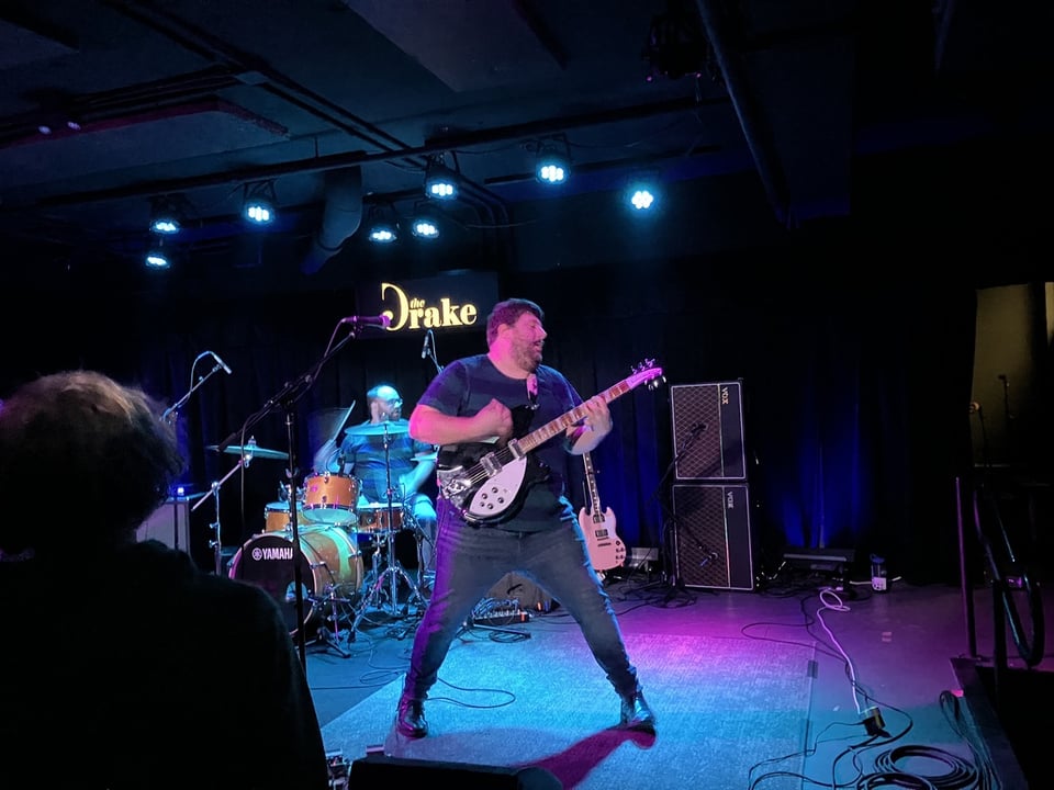 A man with short hair and a few days' stubble plays a Rickenbacker guitar in front of a balding man with glasses on drums.