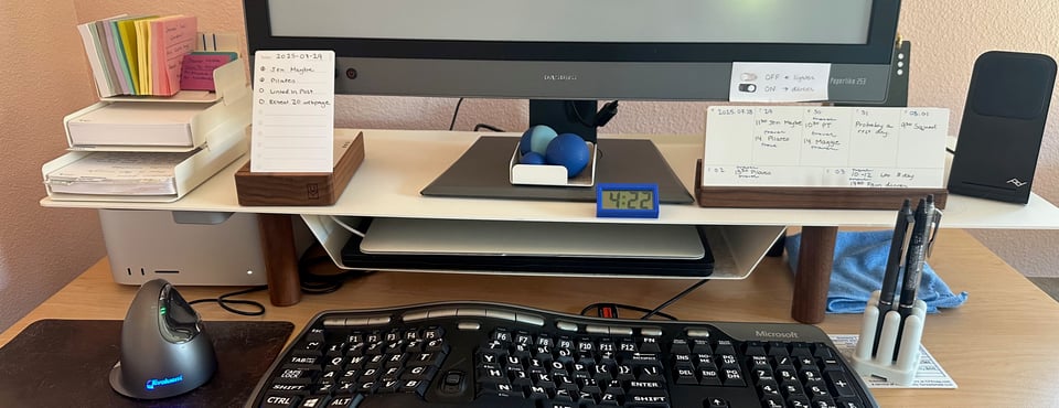 A photo of a tidy desk with a white metal organizer/monitor riser spanning the back edge. From left to right across the top of the riser, there's an index card and post it note organizer with the reverse outline cards shown earlier; a daily to do list; the monitor; a set of MELT balls; a large digital clock; a display of the weekly cards pictured earlier; and a phone charger. In front of the riser, there is an ergonomic mouse, ergonomic keyboard, and pen holder holding the Pilot Frixion pens mentioned elsewhere. You'd correctly infer the person who uses this desk mouses left handed and writes right handed.