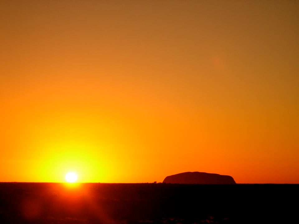 The sky, taking the top three quarters of the photo is burnt orange. At the bottom the horizon is all black in shadow as the sun sets on the left hand side of the picture and Uluru stands next to it on the right hand side.
