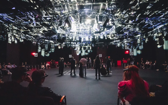 The set-up for Kagami at The Shed, a New York venue. Black curtains encircle the space; audience members are seated in chairs encircling the space, and are standing in front of an area in the centre of the room marked off with white tape. The audience members are all wearing Magic Leap Twos. A large lighting rig hangs overhead.