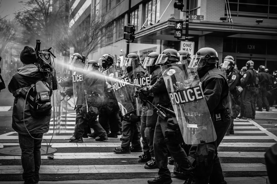 A police line with riot shields and helmets blocks a crosswalk. They are spraying bear spray directed either at a camera person filming the stand off or toward people out of frame on the left side of the photo.
