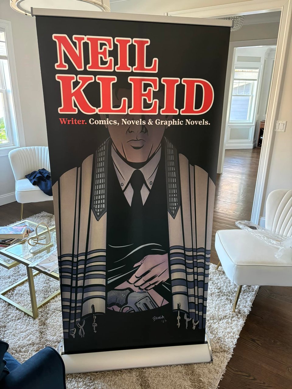 an image of a convention banner stand in a well lit sitting room, showing artwork of a jewish man wrapped in a prayer shawl, pulling up his waistband to reveal a gun, and the words "Neil Kleid" at the top in red and white