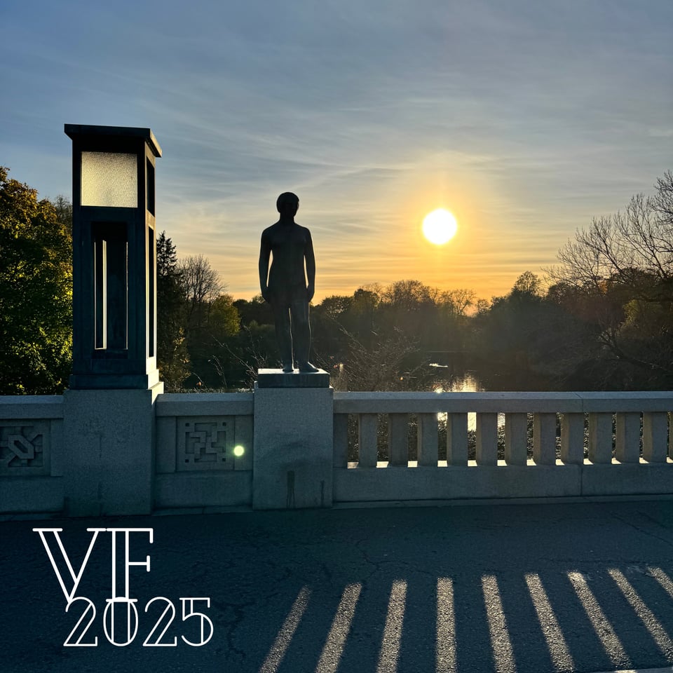 A sidewalk with a stone halfwall at sunest. Atop the halfwall is a statue of man while the sun hangs low to the wooded horizon in a park.