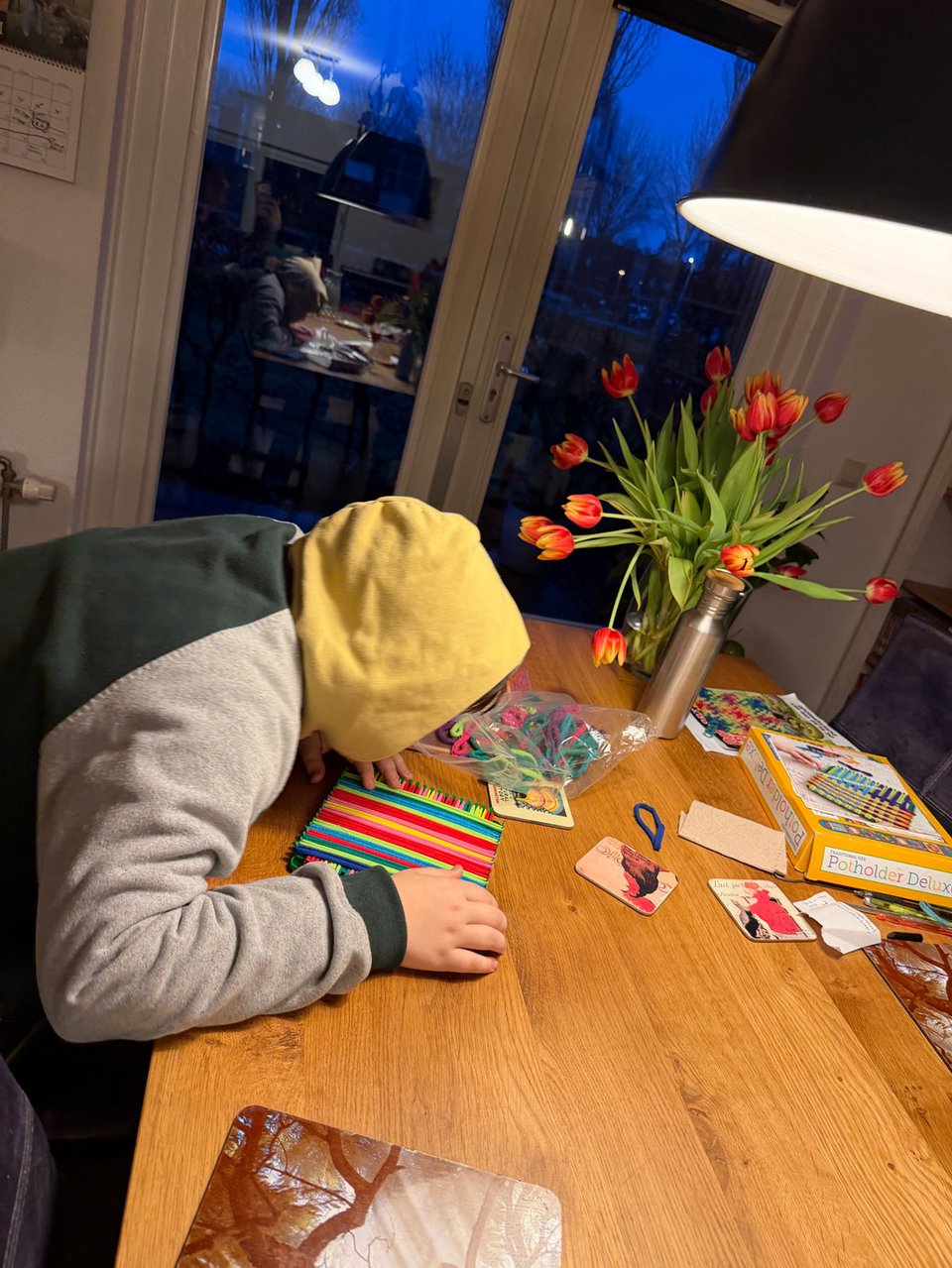 A boy leans over a dining room table as he makes a potholder on a square loom.