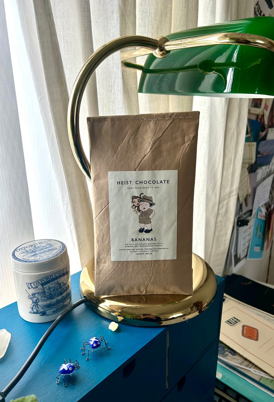 A hand stitched brown paper package containing Heist Bananas chocolate is propped against a brass desk lamp with a green glass shade. There are notebooks scattered behind, and a ceramic pot with 'Oxford Cheese' printed next to the lamp base, plus two tiny glass insects with Scottish flags painted on them. Image by Rowan Ambrose.