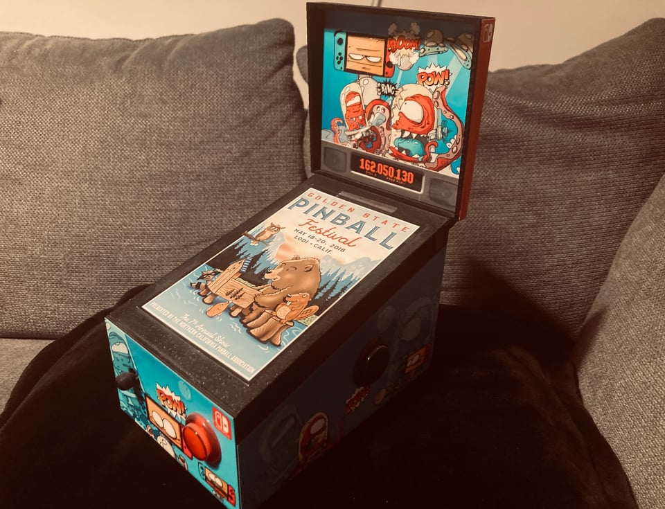 A small pinball cabinet for the Switch console, sitting on a black cushion on a sofa.