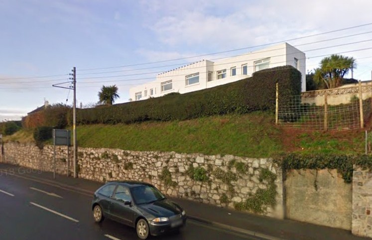 A white rendered pair of semi-detached villas, just visible over a thick box fence. The nearer of the two reveals a curved balcony. The shared middle parapet has a simple decorative pattern of very short fins, almost like crenellations, on it.