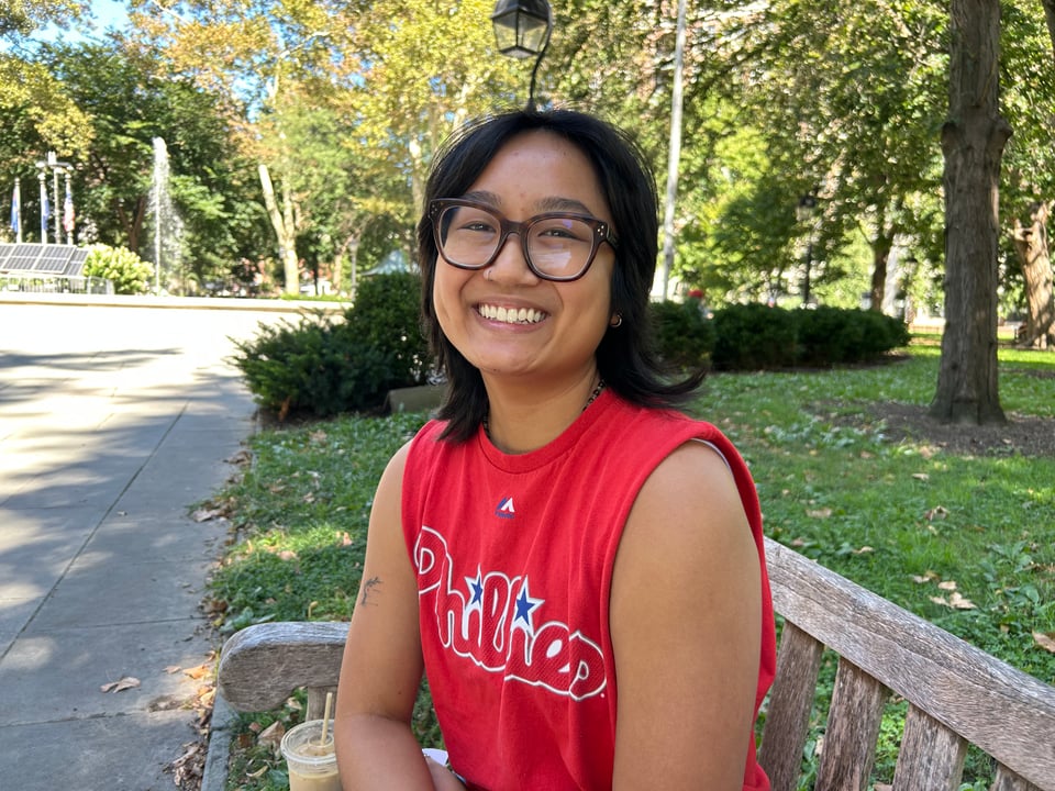 Young Asian person, Samantha, in a Phillies tank top, sitting on a bench at Philadelphia's Washington Square Park, smiling at the camera.