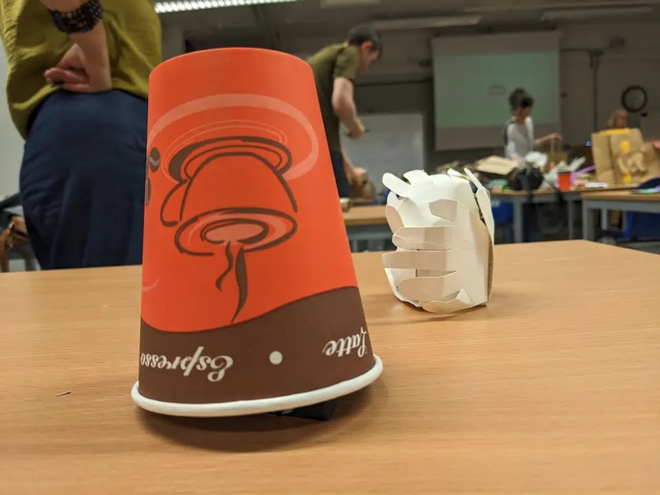 A paper cup is upside down with a gap on one side, and there is a paper sculpture made out of folded hands.