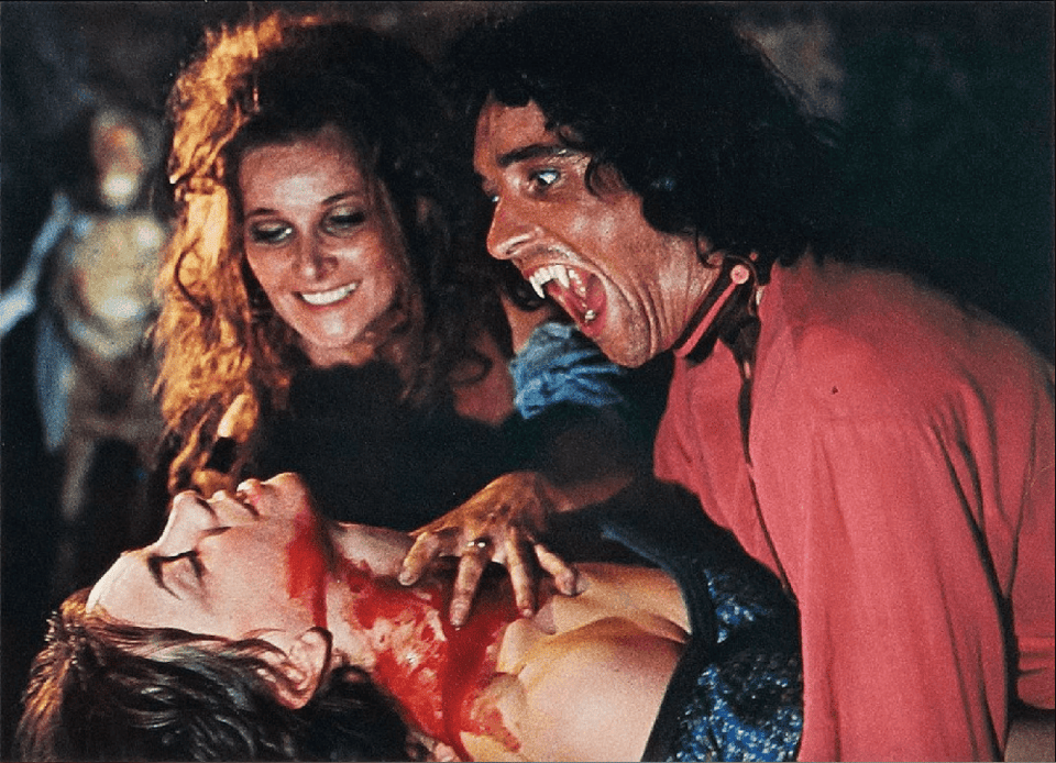 A young woman with a blood covered throat reclines as an older smiling woman and a particularly toothy vampire guy in a red shirt lean over her