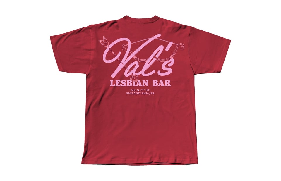 A red t-shirt with "Val's Lesbian Bar" printed in pink script, with a cupid's bow-and-arrow logo resting on the letters, and the address printed below.