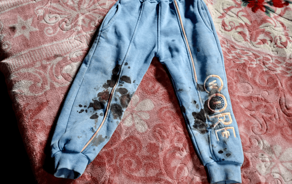 A child's light blue pants laid out on a bed. The pants appear to be stained with blood around the knees and ankles