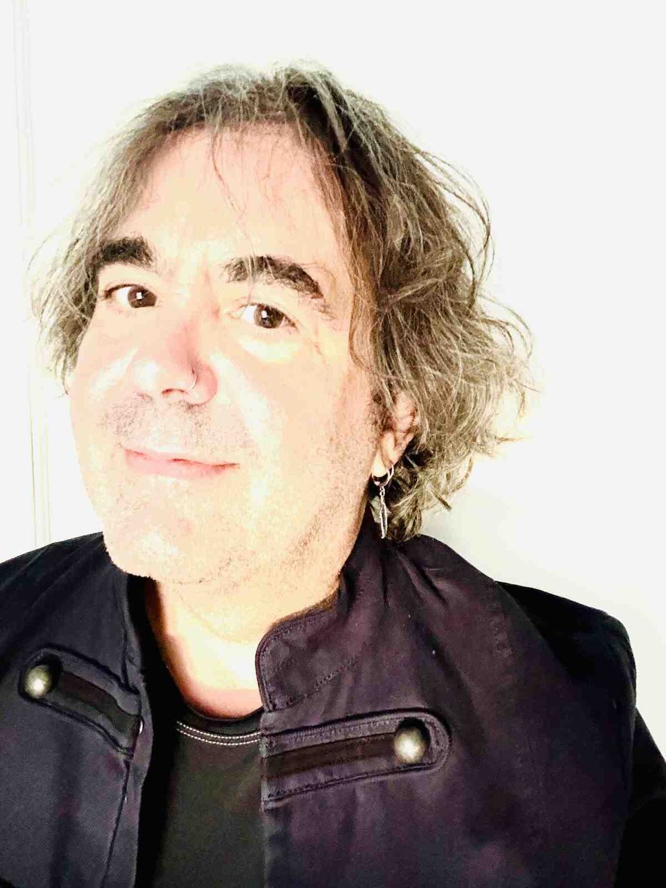 A portrait of a man wearing a black t-shirt and jacket, with a nose ring and blondish-brown hair, smiling at the camera.