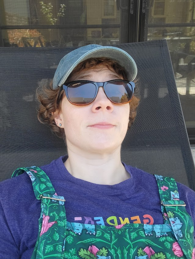 Selfie of a white person with short curly brown hair, reclined in a black lounge chair while wearing sunglasses, a slight smile, a blue baseball cap, a purple shirt that says "GENDER" in rainbow letters, and green bulbasaur overalls.