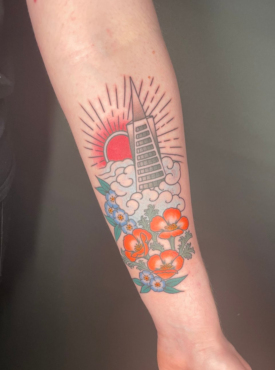 A white person's inner left forearm with a fresh tattoo. It depicts the Transamerica Pyramid with a red sun behind it and a cloud of pale fog swirling at the base, with California poppies and forget-me-nots beneath the fog.