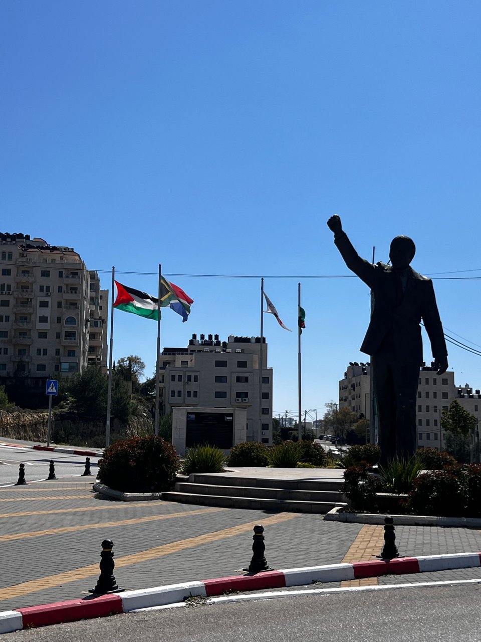 A large status of Nelson Mandela with his fist raised, against a blue sky. Four flags flying behind, including South Africa, Palestine, and the Municipality of Ramallah