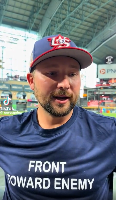 Screenshot of a video from the TikTok account for Our Esquina, a Latine-focused sports outfit. A bearded white man (Seattle Mariners catcher Cal Raleigh) wearing a US baseball cap and a T-shirt that says "Front Toward Enemy" looks down and to his left while answering a question.