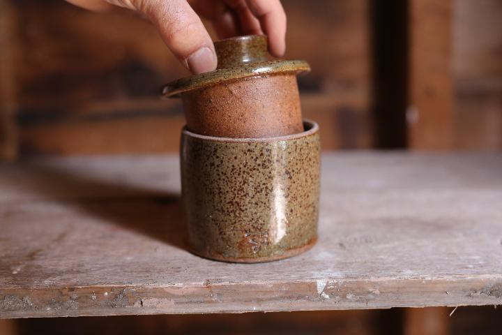 Image of a hand lifting the lid off a butter belle, a brown ceramic pot with lid, showing deep insert of the lid to create a well for the butter. The design is clean, simple and utilitarian but attractive.