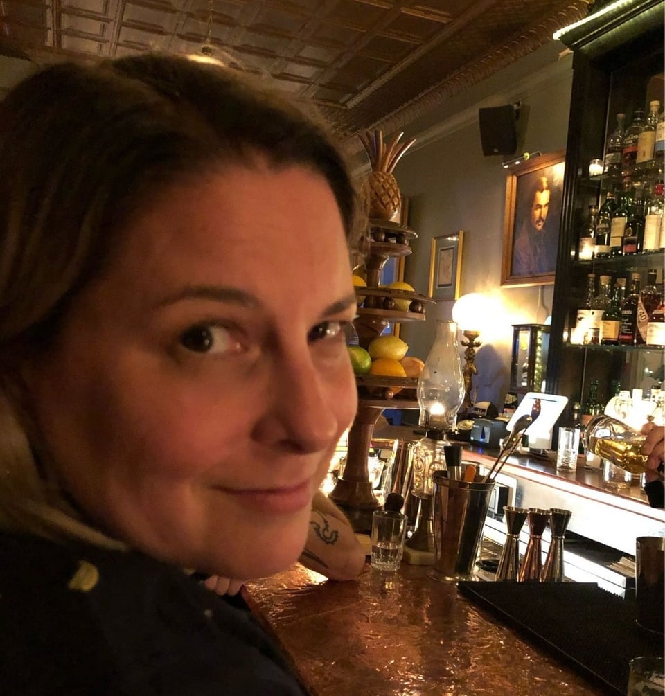 A close up of a woman's face in three-quarter profile. She has a subtle smile, and is seated at a bar, which is visible in the background.