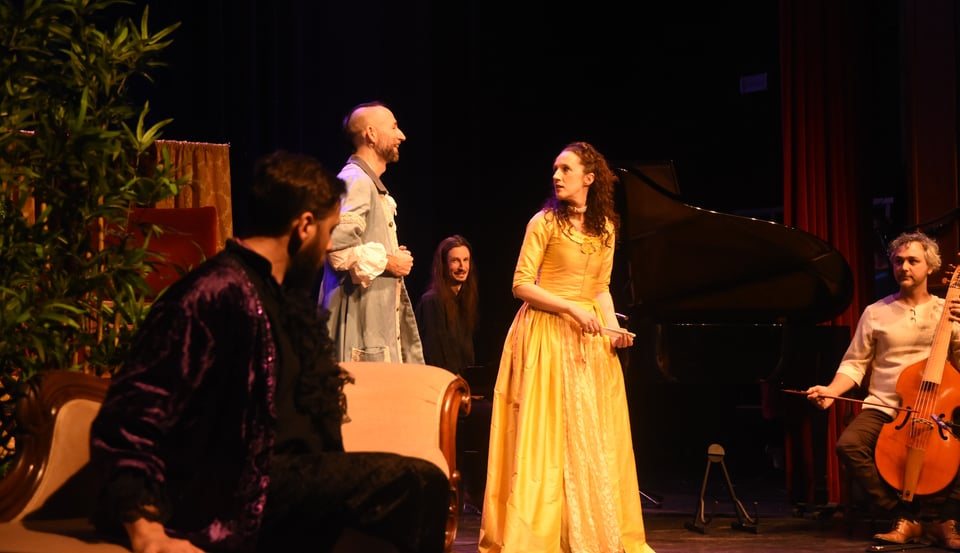 Multiple actors in period drama costumes in a set, accompanied by a cellist and a grand-piano player.