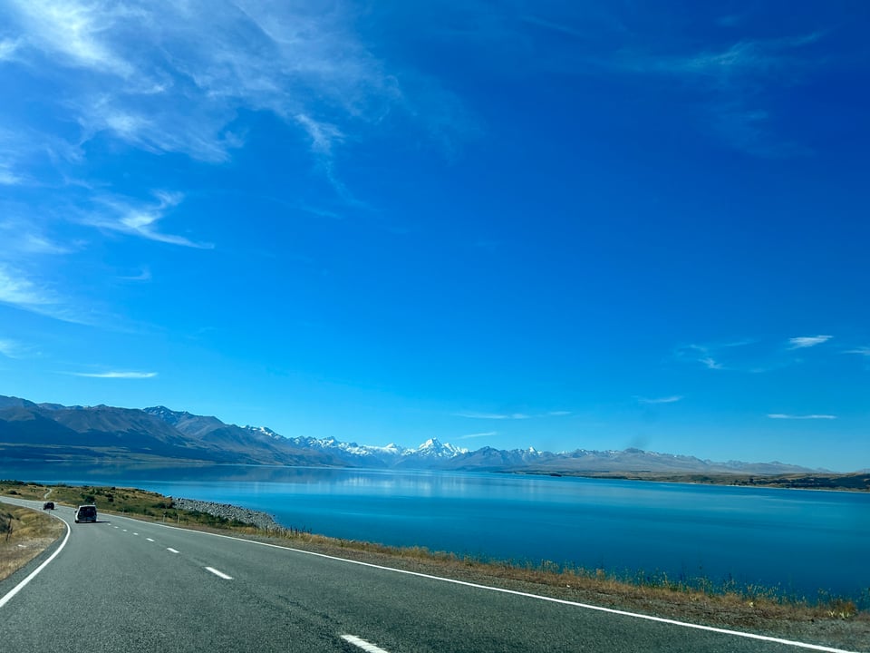 Taken from a car, with the road in the foreground and a vast blue lake with snow-capped mountains under a blue sky in the background.