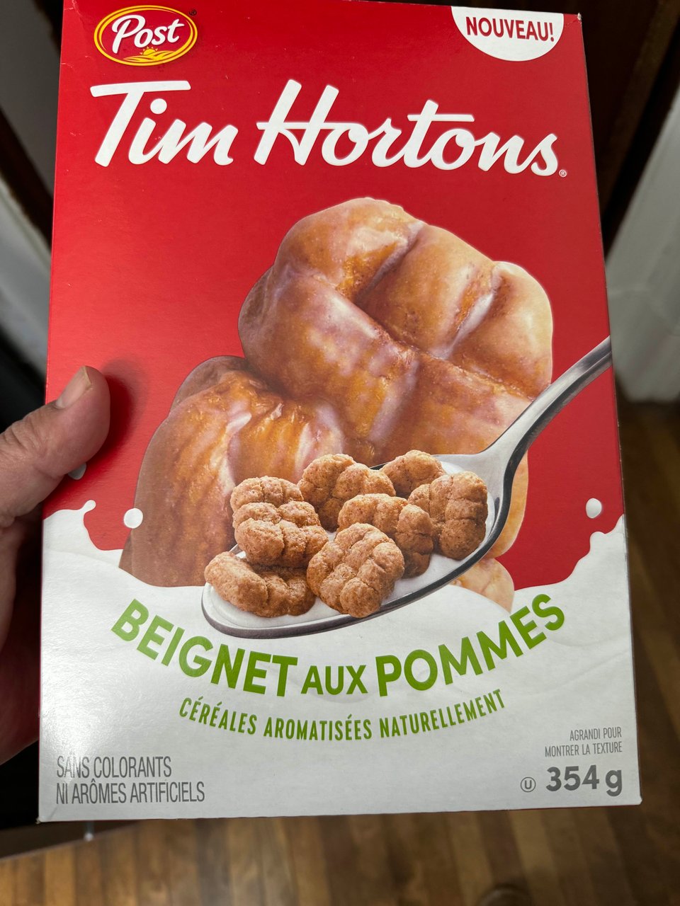 A box of cereal is held before the camera. It's Tim Horton's new Apple Fritter cereal with a spoonful of cereal in front of a large image of an Apple Fritter.