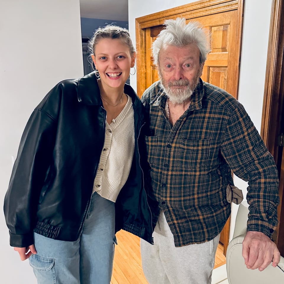 Photo of a young white woman in a black jacket & jeans standing beside her grandfather, whose hair is a mess. They're both smiling
