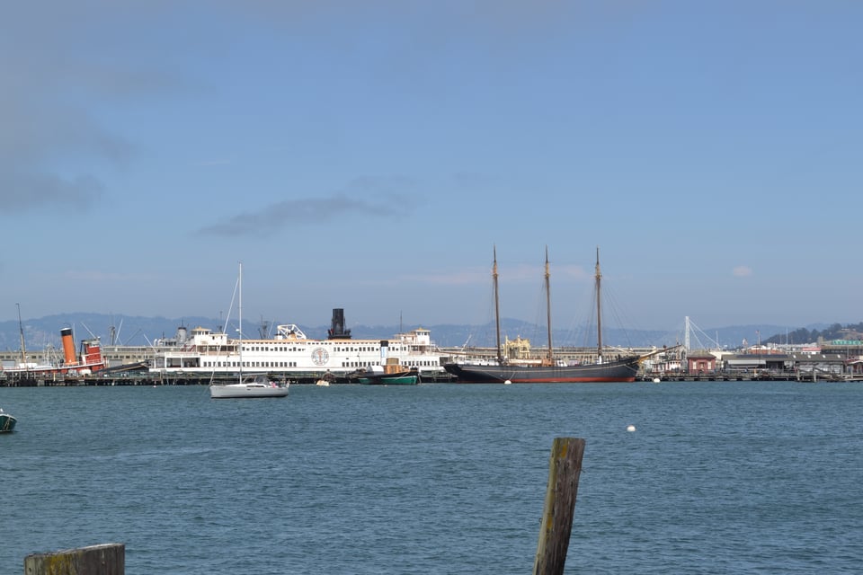 A view across a blue harbor. On the far side is a large ship with a big funnel, a three master, and a small sailing yacht in front of the large ship. Hills can be seen in the distance.
