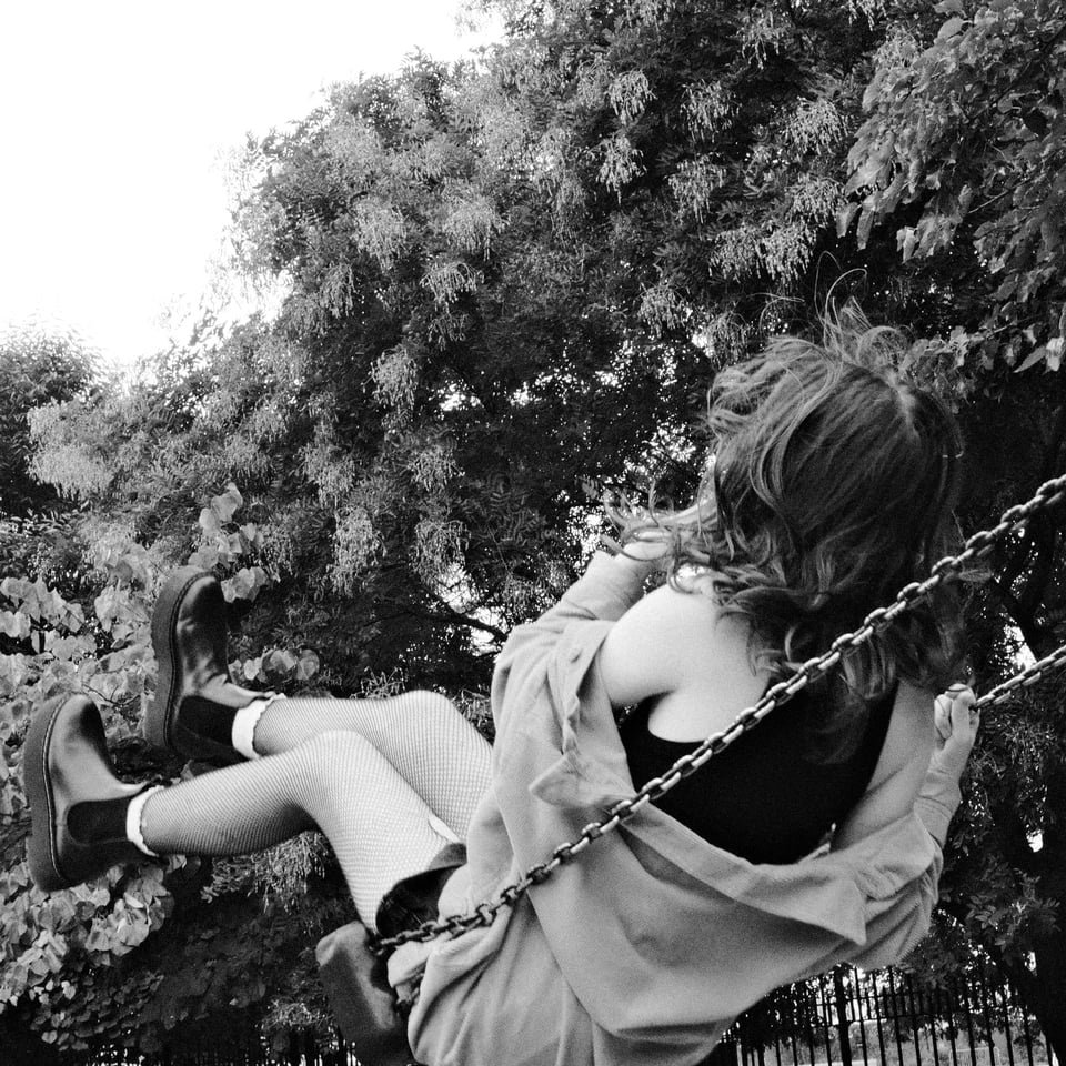 Black and white photograph of Copeland swinging on a swingset, facing away from the camera. Photo taken by Vivienne Altagracia Sanchez