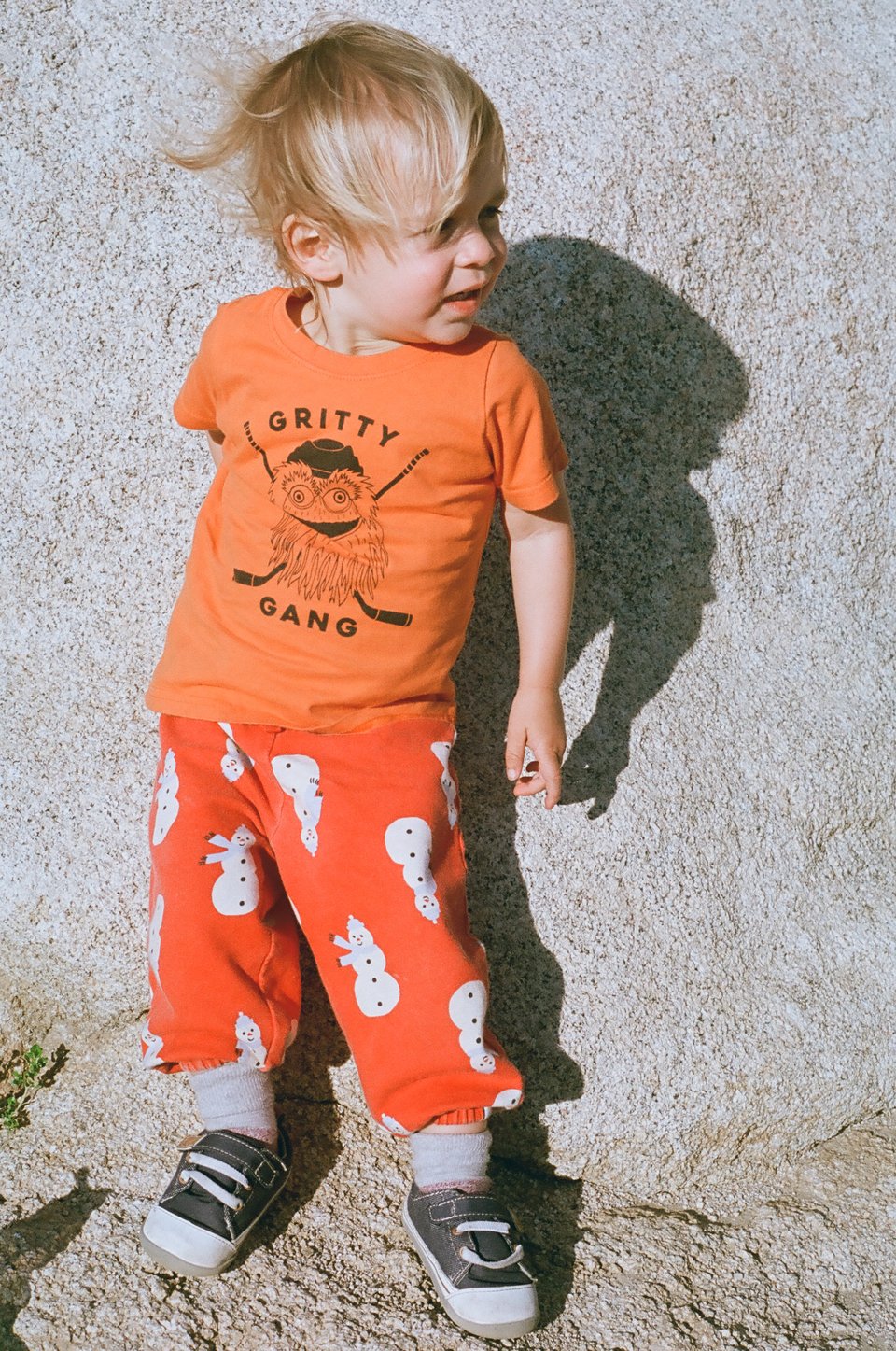 A toddler in an orange "gritty gang" shirt and red pants patterned with snowmen leans against a beige rock in bright sun