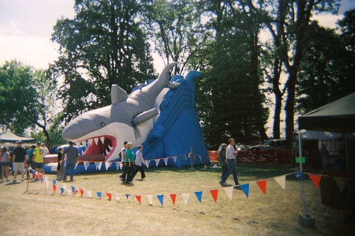 A photograph of a giant inflatable slide that is shaped like a shark riding a wave, so one exits via the shark's mouth. People with out of focus faces wander past, and the bunting and pop-up canopies around indicate it is a fair environment. Lens distortion around the edges forces the eye to focus near the centre, where the shark's confused eye is.