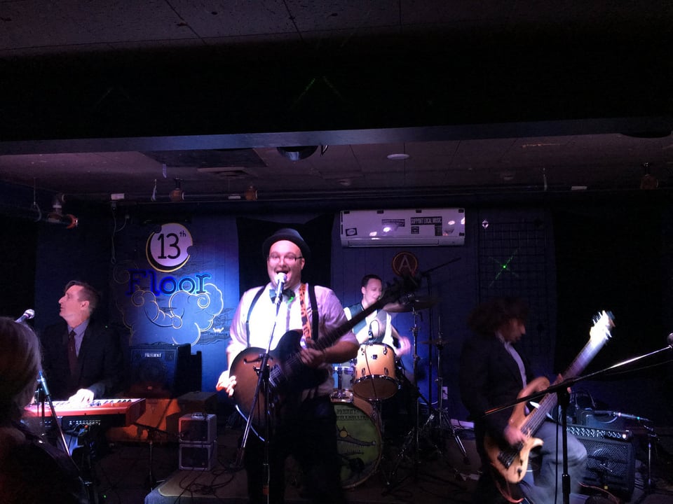 Four musicians -- a guitarist front and center, a keyboard player to his right, a bass player to his left and a drummer behind him -- play onstage.