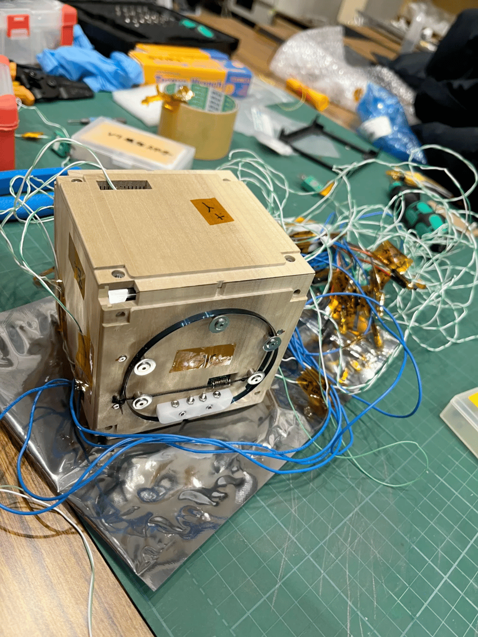 A photograph of a workbench covered with a green cutting mat. On the cutting mat sits a tangle of wires protruding from a cube made of light wood that appears to be pine. The cube is bolted together with screws in each corner of the cube. The side of that faces us has five large shiny screws embedded, and they support a black ring of plastic, though it's not clear what the function of this ring is. The cube is also sitting on top of what appears to be a shiny static bag. In the back are various electronic components in a roll of tape, and sundry tools.