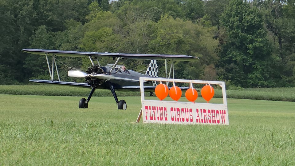 A vintage biplane taxiing across a grassy airfield, with a white "Flying Circus Airshow" sign holding four orange balloons in the foreground and dense green trees behind.