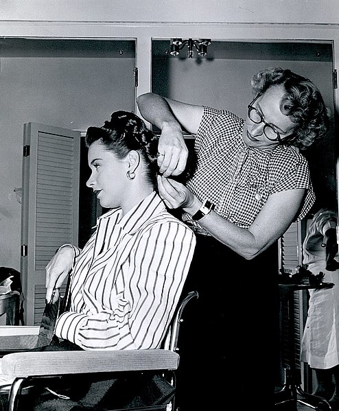 A woman sites in a salon chair, another woman styles her hair.