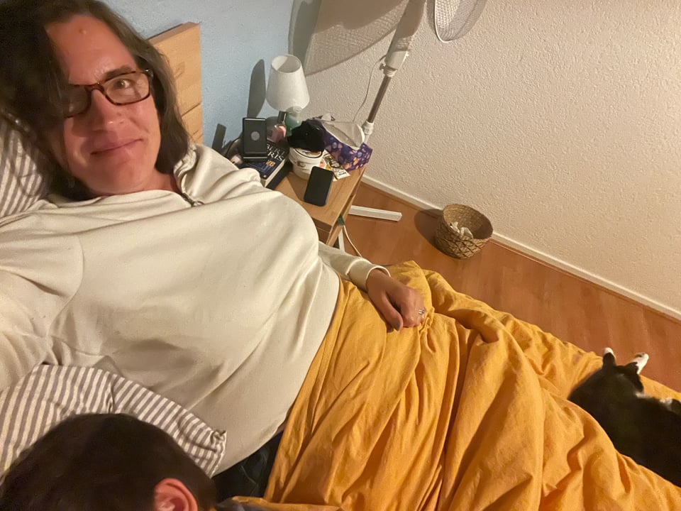 A woman looks at the camera and is snuggled by a cat on one side and a sleeping child on the other.