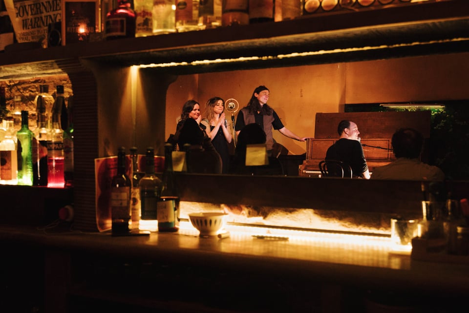 Sienna Sears, Sydney Shepherd, and Adrian Enscoe look on with pride/emotion as Christopher Sears looks out at the crowd from the piano. The whole group are reflected in the mirror of the bar.