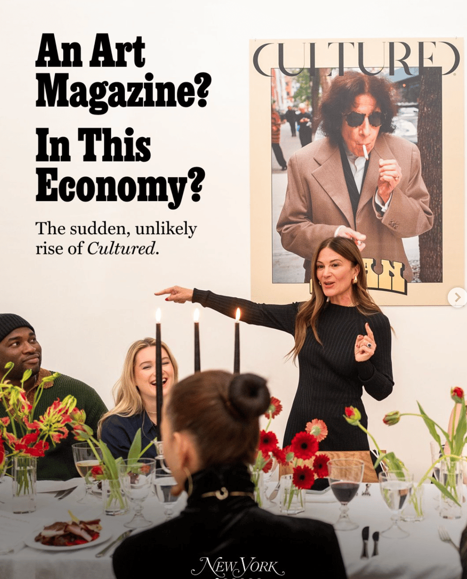 CULTURED Mag EIC and founder Sarah Harrelson stands at long dinner table with seated guests and flower arrangements, extending her arm out as though pointing to someone out-of-frame. Behind her is a poster-sized reproduction of a magazine cover featuring Fran Leibowitz