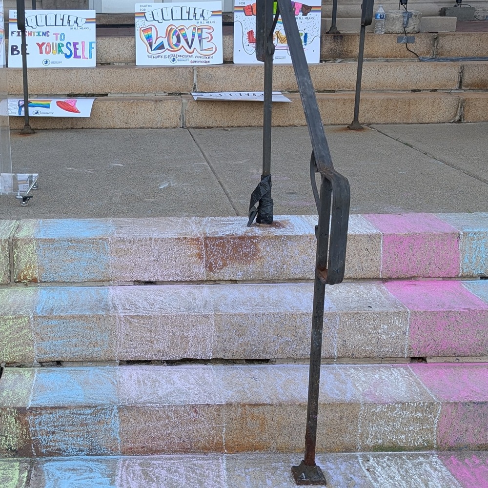 Steps with a railing up the middle are chalked in the colors of the pride and trans flags. Resting on a higher set of steps are signs reading Equality is fighting to be yourself and Equality is Love drawn in a rainbow of colors