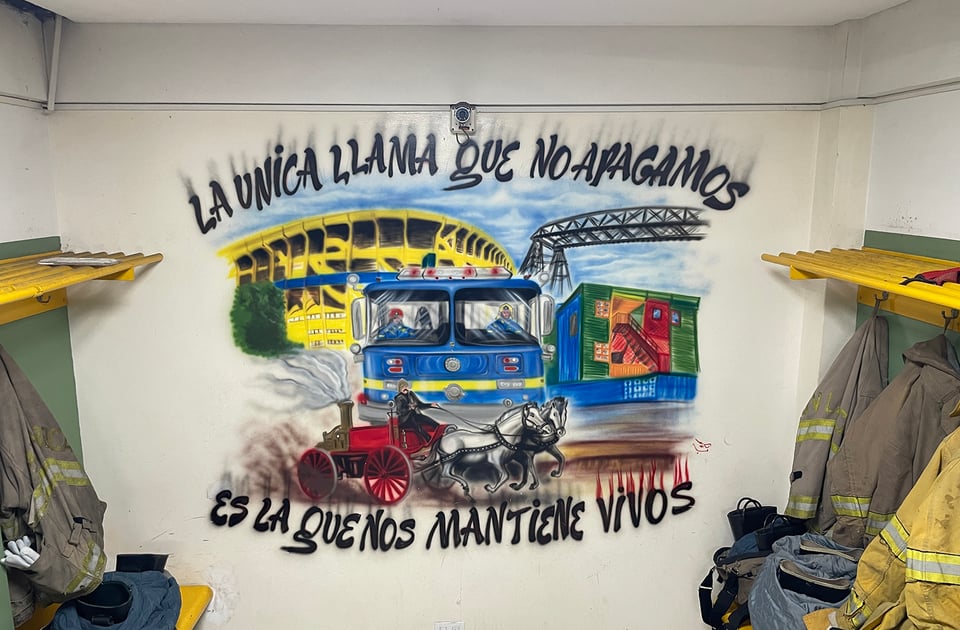 A mural depicts firefighters from the neighborhood of La Boca with various touristic highlights of the neighborhood, the famous bridge, the football stadium, the colorful tenement housing and the old fire engine pump being pulled by white horses.