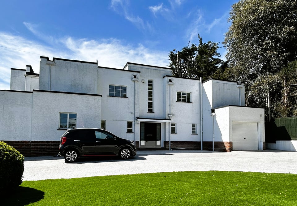 a large white 1930s building with a small black car parked outside. The building is broadly symmetrical, with a central entrance door and double height crittell window. Either side are more windows, then an integral garage and another block.