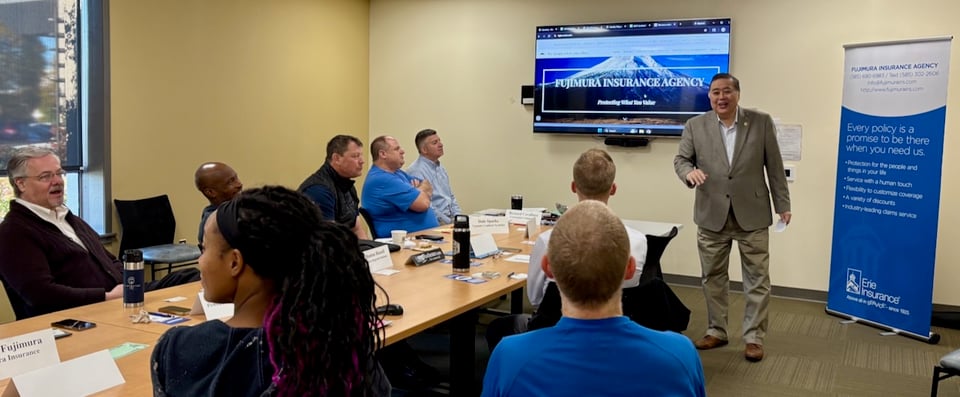 Robert smiles as he presents to seated NRG members about Fujimura Insurance Agency, with his website on a wall-mounted display and a retractable vertical banner highlighting his business.
