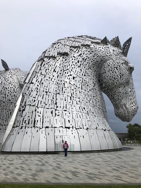 Me, looking very tiny, standing beside a 98-foot tall metal sculpture of a horse head. The sculpture is matte-silver in color and made up of hundreds of separate pieces that give a kind of digital lattice effect. A second, equally massive horse head is behind. We are all on a gray stone-flagged plaza beneath a cloudy sky