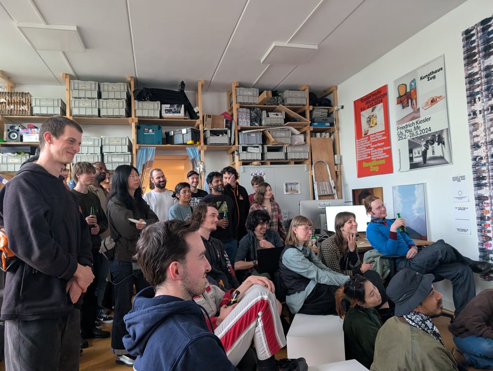 An audience of people, some seated, some standing, watch a screen that is off-frame.