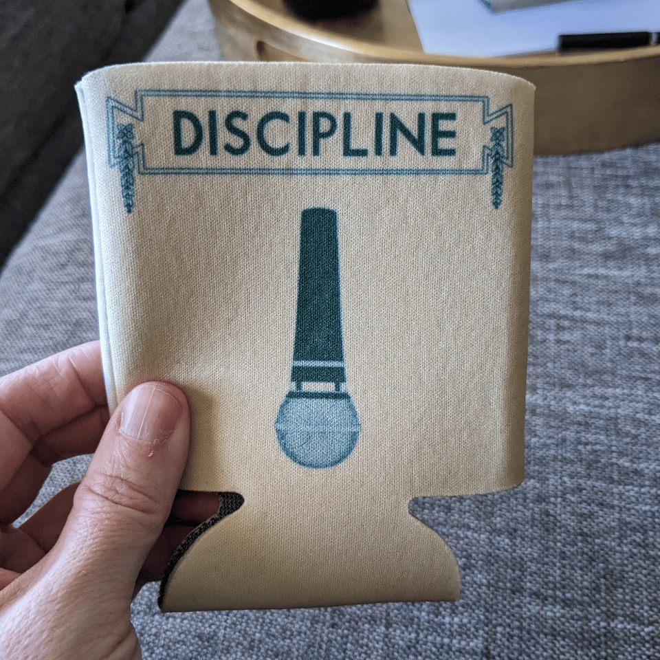 A hand holding a pail beige coozie with evergreen printed image and text. The image is an upside-down microphone. The text above it says DISCIPLINE in all caps with a box around it and some greenery like ivy hanging off the box.