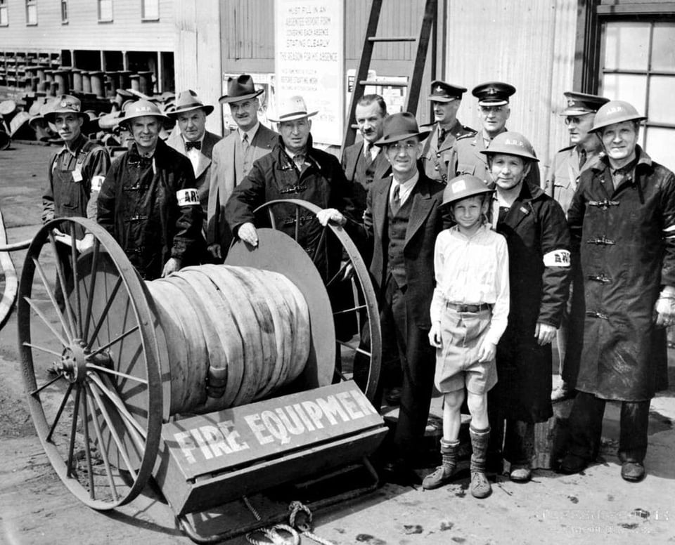 Black and white photo. A group of men and one boy wearing military and civilian hats stand in front of a fire hose on wheels. Some are wearing armbands that read "ARP"