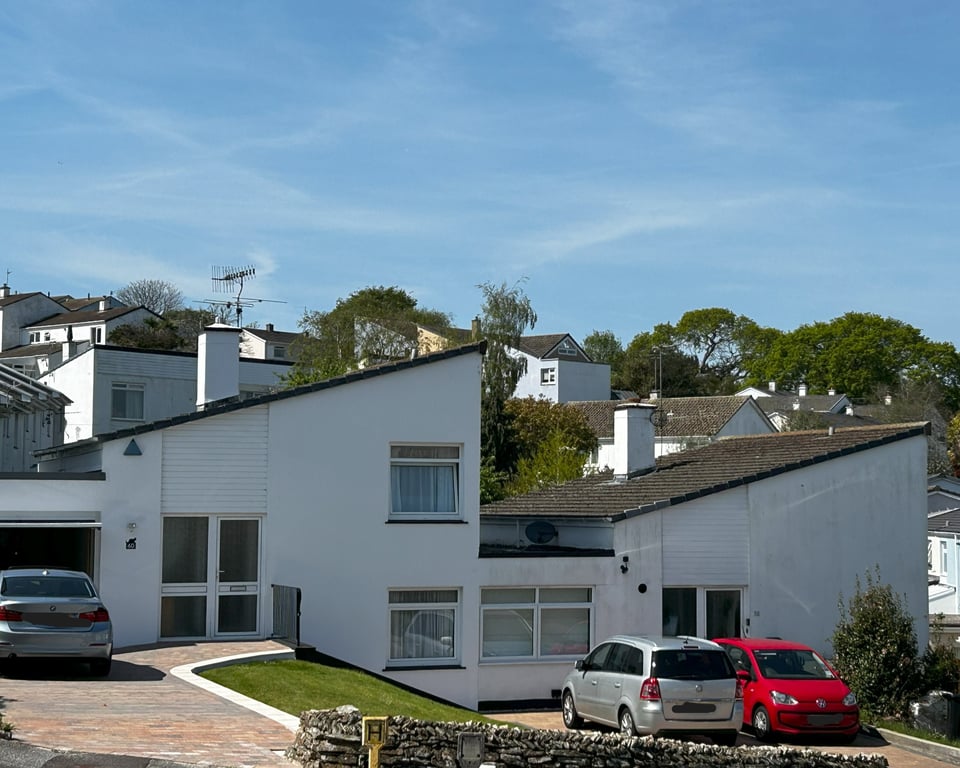 In the foreground are two white rendered houses. Each is split storey, so from the front door you would go up or down stairs to get to the main rooms. Each has a big mono-pitched roof and a link garage. One is still using their garage, the other has filled that in to create more rooms. on the hillside above there are more buildings in the same style.